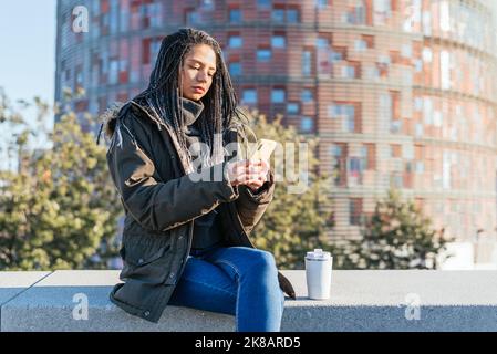 Femme hispanique ciblée avec messagerie texte « Dreadlocks » sur un téléphone portable, assise sur un banc avec thermos de café chaud en ville Banque D'Images