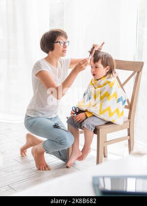 Mère coupe les cheveux de son fils par elle-même. Le petit garçon est assis, recouvert de tissu, et tient une paire de ciseaux. Nouveau normal en cas de coronavirus COVID-19 qua Banque D'Images