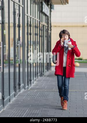 Femme en duffle-coat rouge parle par smartphone. Une femme descend dans la rue en prenant une tasse de café à emporter et en prenant un chèque papier. Banque D'Images