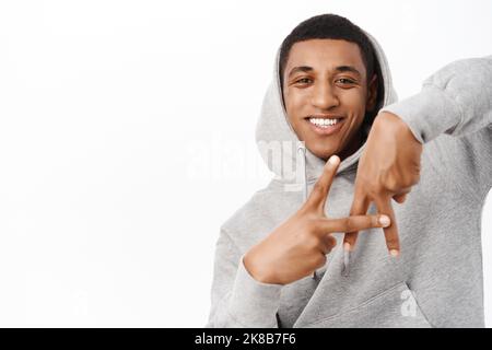 Un homme afro-américain de bonne qualité montre LE geste DE LA main, debout dans un sweat à capuche gris sur fond blanc Banque D'Images