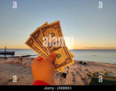 Un homme tenant trois billets d'un dollar dans sa main sur fond de mer, de plage de sable et de ciel bleu le jour d'été ensoleillé. Le concept de l'argent, de la finance, de la monnaie, des voyages, du tourisme, vacances, repos Banque D'Images