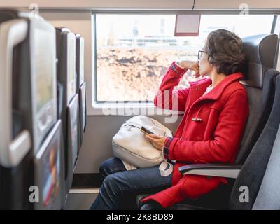 Femme souriante en duffle-coat rouge avec sur smartphone se trouve près de la fenêtre dans le train de banlieue. Voyager en véhicule terrestre. Banque D'Images