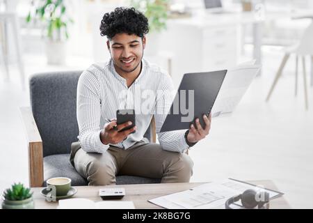 Un jeune homme d'affaires utilisant un téléphone portable tout en passant par des documents administratifs dans un bureau. Banque D'Images