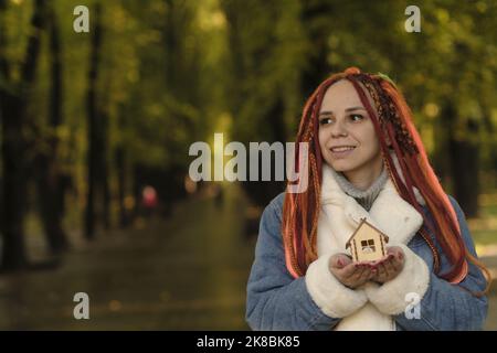 Bonne femme tenant le modèle à la maison dans le parc. Jeune femme positive avec des dreadlocks tenant une petite maison en bois souriant et regardant la caméra dans le parc d'automne. Banque D'Images