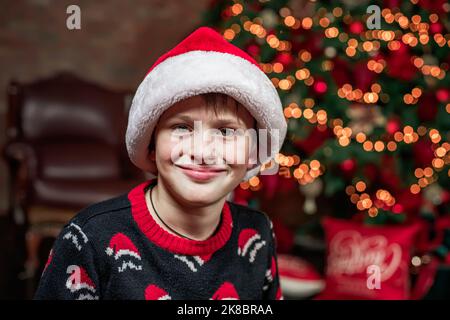 Un garçon dans le chapeau du Père Noël dans le fond des lumières de l'arbre de Noël. La veille de Noël, un garçon en attente de cadeaux. Banque D'Images