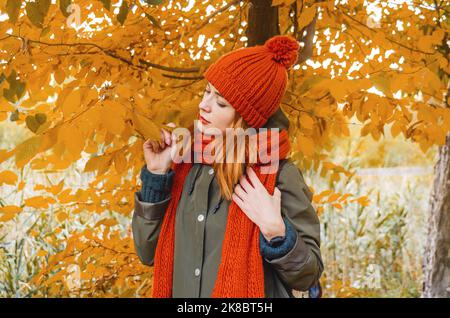 La jeune femme se tient sous l'arbre avec des feuilles jaunes. Vêtu d'un bonnet et d'une écharpe tricotés orange tendance. Ambiance d'automne. L'émotion de la tristesse. Banque D'Images