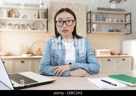 Consultation en ligne d'un psychologue. Une jeune femme asiatique psychologue spécialisée effectue des rendez-vous de patients en ligne. Assis à la table à la maison avec un ordinateur portable, bloc-notes et regardant la caméra. Banque D'Images