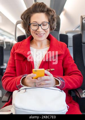 Femme souriante dans un duffle-coat rouge envoyant un SMS sur un smartphone dans un train de banlieue. Voyager en véhicule terrestre. Banque D'Images