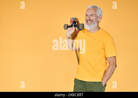 Patineuse d'homme ancien à barbe heureuse tenant une planche à roulettes isolée sur jaune. Banque D'Images