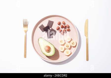 Avocat, banane, morceaux de chocolat et huzelnuts sur une assiette avec fourchette et couteau. Ingrédients pour smoothie. Vue de dessus, plan d'appartement. Banque D'Images