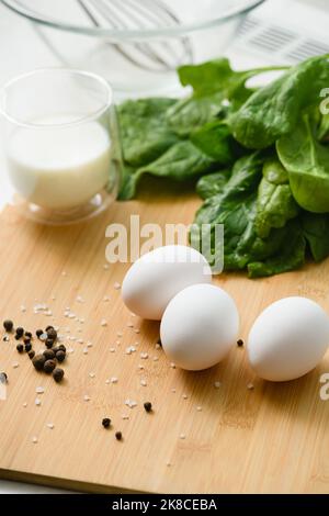 Trois œufs de poulet blancs sur une planche à découper avec un bouquet d'épinards frais. Un verre de lait en arrière-plan. Banque D'Images