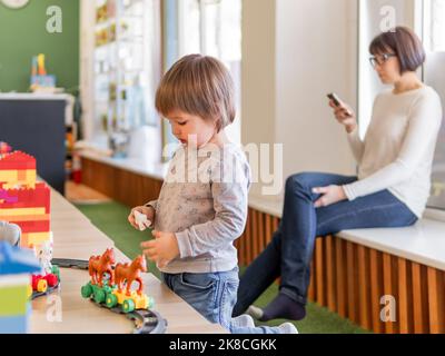 Tout-petit joue avec des blocs de jouets colorés tandis que sa mère ou sa gardienne d'enfants textent dans un smartphone. Un petit garçon se trouve sur le constructeur du jouet. Jardin d'enfants. Banque D'Images