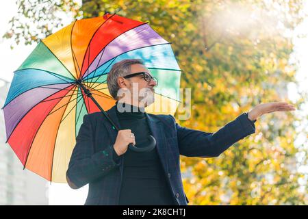 Un homme souriant, d'âge moyen, barbu, aux cheveux gris dans un blazer et un col roulé tenant un parapluie arc-en-ciel vif sur sa tête, en vérifiant si son pluie avec sa main. Arbres flous au soleil. Photo de haute qualité Banque D'Images