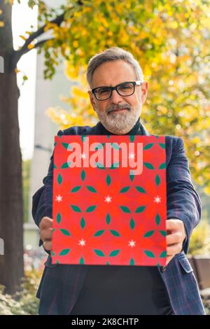 Un homme mûr souriant, positif, barbu, aux cheveux gris avec des lunettes, tenant un sac cadeau rouge, le montrant à la caméra dans le parc. Photo de haute qualité Banque D'Images