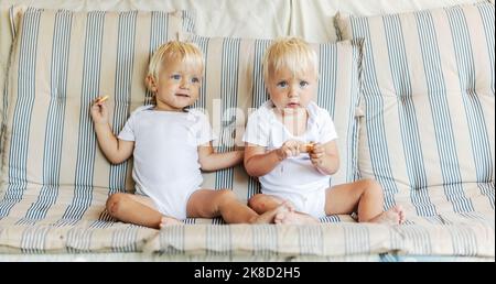 Adorables enfants identiques avec la lumière entendre et les yeux bleu ciel. Les jumeaux de bébé blancs s'assoient sur un confortable canapé beige dans le salon Banque D'Images