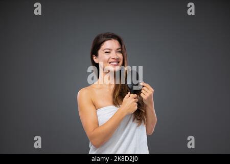 Femme belle et bien entretenue. Une femme enveloppée d'une serviette blanche autour de son corps se tient devant un mur gris et utilise une brosse à cheveux après la douche Banque D'Images