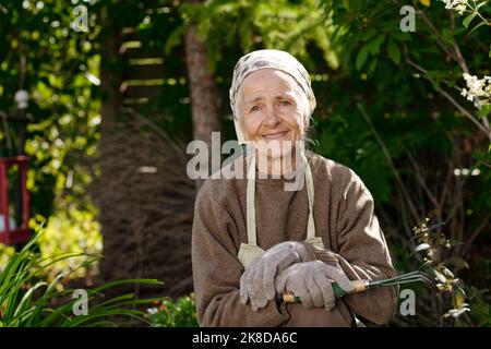 Bonne femme âgée avec petit râteau pour desserrer le sol regardant la caméra tout en se tenant dans le jardin avec des plantes vertes le jour ensoleillé Banque D'Images