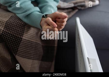 Une fille chauffe les mains gelées au-dessus du radiateur chaud, en gros plan. Femme portant un chandail en laine se réchauffant tout en étant assise près d'un radiateur de chauffage. Femme Banque D'Images