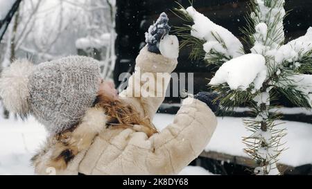 Décoration extérieure arbre de Noël. Cutie, jolie petite fille décore l'arbre de Noël recouvert de neige avec des jouets de glace faits maison, en chute de neige, dans la forêt, près de la vieille maison en bois. Happy time sur la neige hiver. Activités familiales en plein air. Ralenti. Photo de haute qualité Banque D'Images