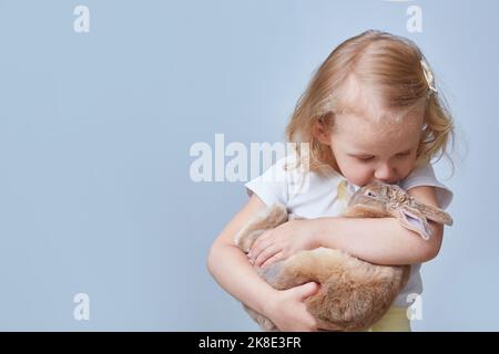 Une jolie fille embrasse et embrasse un lapin. Copier l'espace sur un arrière-plan bleu. Banque D'Images