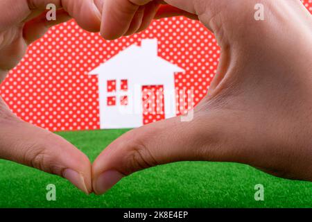 Maison en papier au milieu d'une main en forme de coeur sur un fond blanc-pointillé rouge avec de l'herbe Banque D'Images