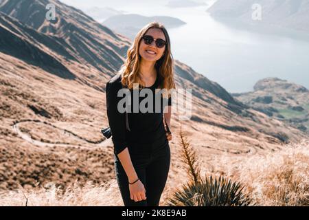 jeune femme caucasienne avec des cheveux en vrac dans des lunettes de soleil et un sac à dos sur son dos sourit à la caméra calme et détendu sur le sommet d'une haute montagne calme Banque D'Images
