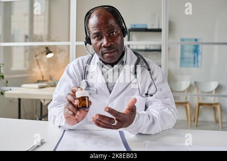 Portrait d'un pédiatre africain mature dans un casque offrant une consultation en ligne sur les pilules tout en étant assis à table au bureau Banque D'Images