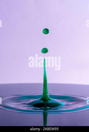 Gros plan de gouttes d'eau de couleur verte éclaboussant sur la surface sur fond violet. Banque D'Images
