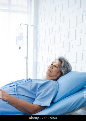 Femme asiatique sénior, cheveux gris, dormant sur le lit dans une chambre d'hôpital blanche moderne, de style vertical. Une vieille dame est malade et allongé sur le lit du patient, Banque D'Images