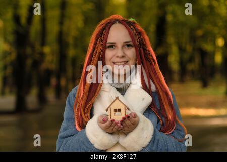 Bonne femme tenant le modèle à la maison dans le parc. Jeune femme positive avec des dreadlocks tenant une petite maison en bois souriant et regardant la caméra dans le parc d'automne. Banque D'Images