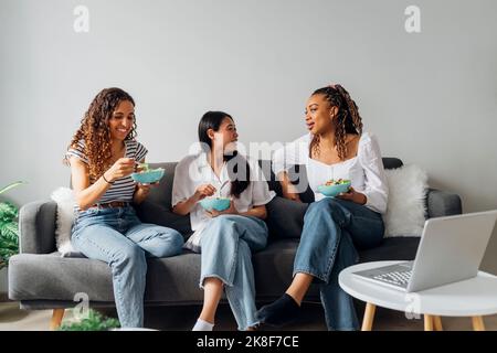 Jeunes colocataires souriants et multiraciaux qui mangent de la salade ensemble, assis sur un canapé à la maison Banque D'Images