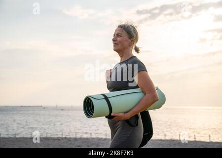 Femme mature tenant un tapis d'exercice au coucher du soleil Banque D'Images