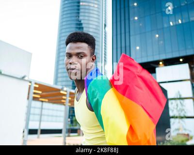 Jeune homme enveloppé dans un drapeau arc-en-ciel par des bâtiments modernes Banque D'Images