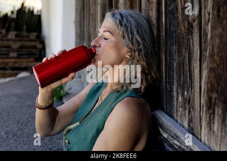 Femme âgée avec les yeux fermés de l'eau potable de la bouteille par porte en bois Banque D'Images