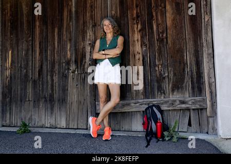Heureuse femme âgée portant des chaussures orange avec les bras croisés penchée sur la vieille porte de la grange en bois Banque D'Images