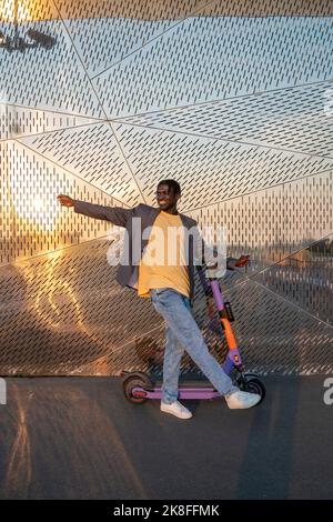 Jeune homme d'affaires souriant avec scooter électrique à pousser devant un mur en métal Banque D'Images