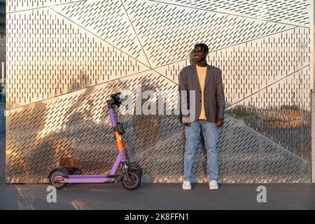 Jeune homme d'affaires souriant debout à bord d'un scooter électrique en face d'un mur en métal Banque D'Images