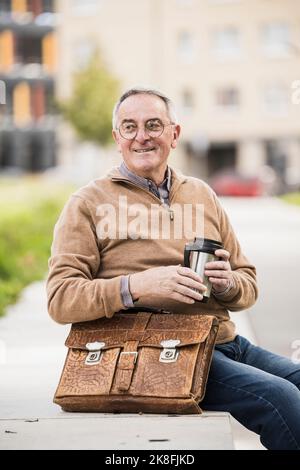 Homme souriant tenant un contenant à boisson isolé assis avec un sac sur le banc Banque D'Images