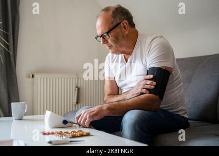 Homme âgé utilisant un appareil de mesure de la tension artérielle assis sur un canapé à la maison Banque D'Images