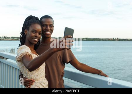 Jeune couple souriant prenant le selfie debout au bord de la rivière Banque D'Images