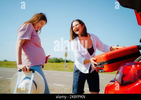 Bonne femme en train de charger sa valise dans le coffre de la voiture et de regarder une amie enceinte Banque D'Images