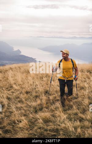 Homme mûr souriant portant un chapeau marchant sur l'herbe avec des bâtons de randonnée Banque D'Images