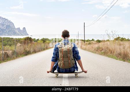 Homme avec sac à dos assis sur une planche à roulettes devant le ciel Banque D'Images