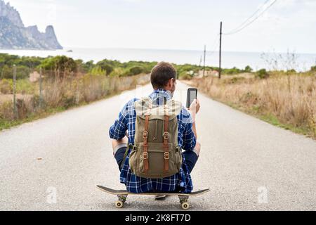 Homme portant un sac à dos avec un smartphone assis sur un skateboard Banque D'Images