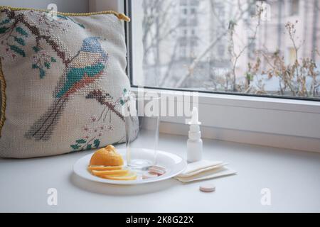 Froid de saison: Une tasse en verre vide, citron en tranches, pilules instantanées et vaporisateur nasal avec serviettes en papier sur le rebord de la fenêtre. À côté du modèle vintage brodé p Banque D'Images