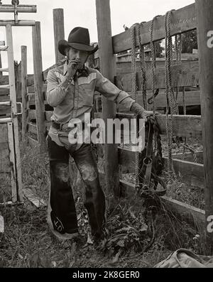 Portrait monochrome vintage d'un cowboy de rodéo portant des bonbonnes de cuir et se penchant contre une chute dans l'écriture sur Stone, Alberta Canada. Vers 1983 Banque D'Images