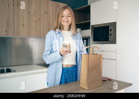 Livraison de nourriture. Jeune femme déballant le sac en papier après avoir fait des achats en utilisant un smartphone dans la cuisine intérieure à la maison, espace de copie. Banque D'Images