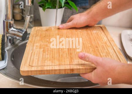 Un homme lave une planche à découper en bambou en bois dans l'évier de cuisine sous l'eau courante. Lavage doux à la main d'une planche à découper en bois. Nettoyage des produits en bois sales de cuisine. Banque D'Images