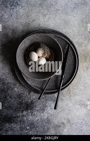 De la table minimaliste ci-dessus pour le dîner de Pâques avec vaisselle sombre et couverts sur les assiettes en céramique et bol avec nid et œufs de caille sur le béton Banque D'Images