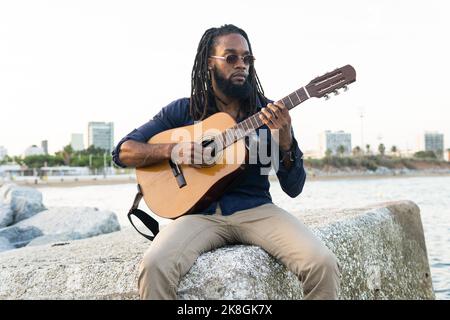 Homme ethnique barbu en lunettes de soleil jouant de la musique sur guitare acoustique tout en étant assis sur la frontière de pierre près de la rivière contre la ville Banque D'Images